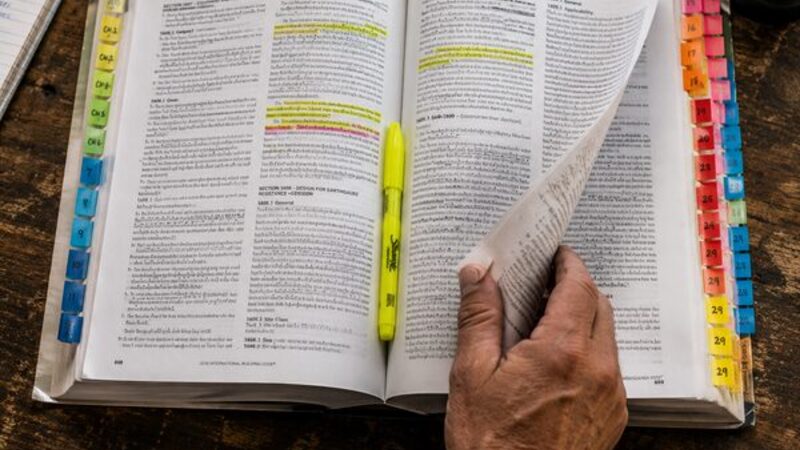 Open International Building Code reference book lined with colored index tabs and highlighted exam-prep passages, contractor's hand turning a page